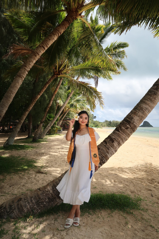 A picture of a girl smiling in a white dress standing on the beach under a palm tree. She is wearing her Phi Theta Kappa gold stole and honor cords, with a white hibiscus flower tucked behind her ear, capturing both her academic achievements and the tropical paradise around her.