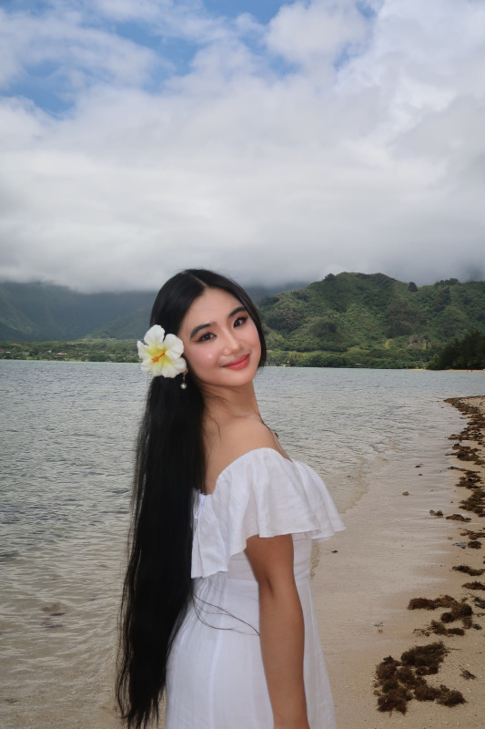 A picture of a girl in a white dress standing on the beach, her side profile visible as she looks over her shoulder behind her, with a white hibiscus flower tucked behind her ear, capturing the gentle ocean breeze and the serene coastal atmosphere.