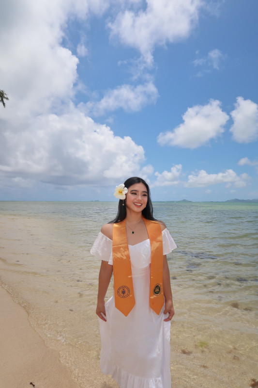 A candid picture of a girl laughing in a white dress standing on the beach, wearing her Phi Theta Kappa National Honor Society gold stole, with a white hibiscus flower tucked behind her ear. She is holding her dress playfully.