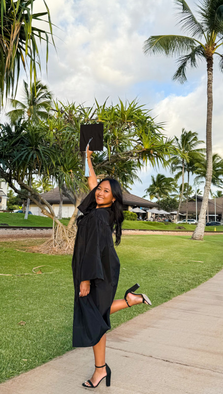 Caydence in her black Leeward Community College gown holding up her black cap and light blue tassel with one leg up.
