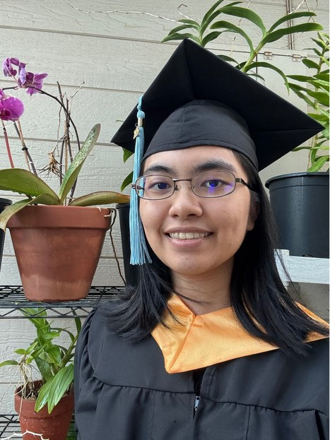 A photo of myself wearing the LEE CC cap, gown, and honors hood. There are potted plants in the backgrond of the photo.