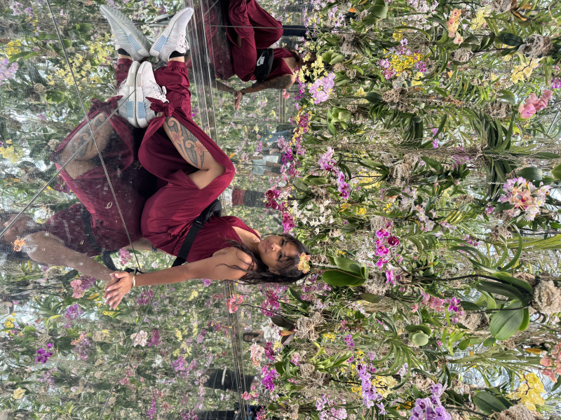 selfie of a woman sitting on a mirror floor, in a maroon jumpsuit that has a slit on the leg, with the leg showing a hawaiian tribal tattoo with plumerias on it, with white shoes. There are beautiful colorful flowers surrounding endlessly hanging all around above her, so low that if she were to stand up, she would be standing in that field of flowers
